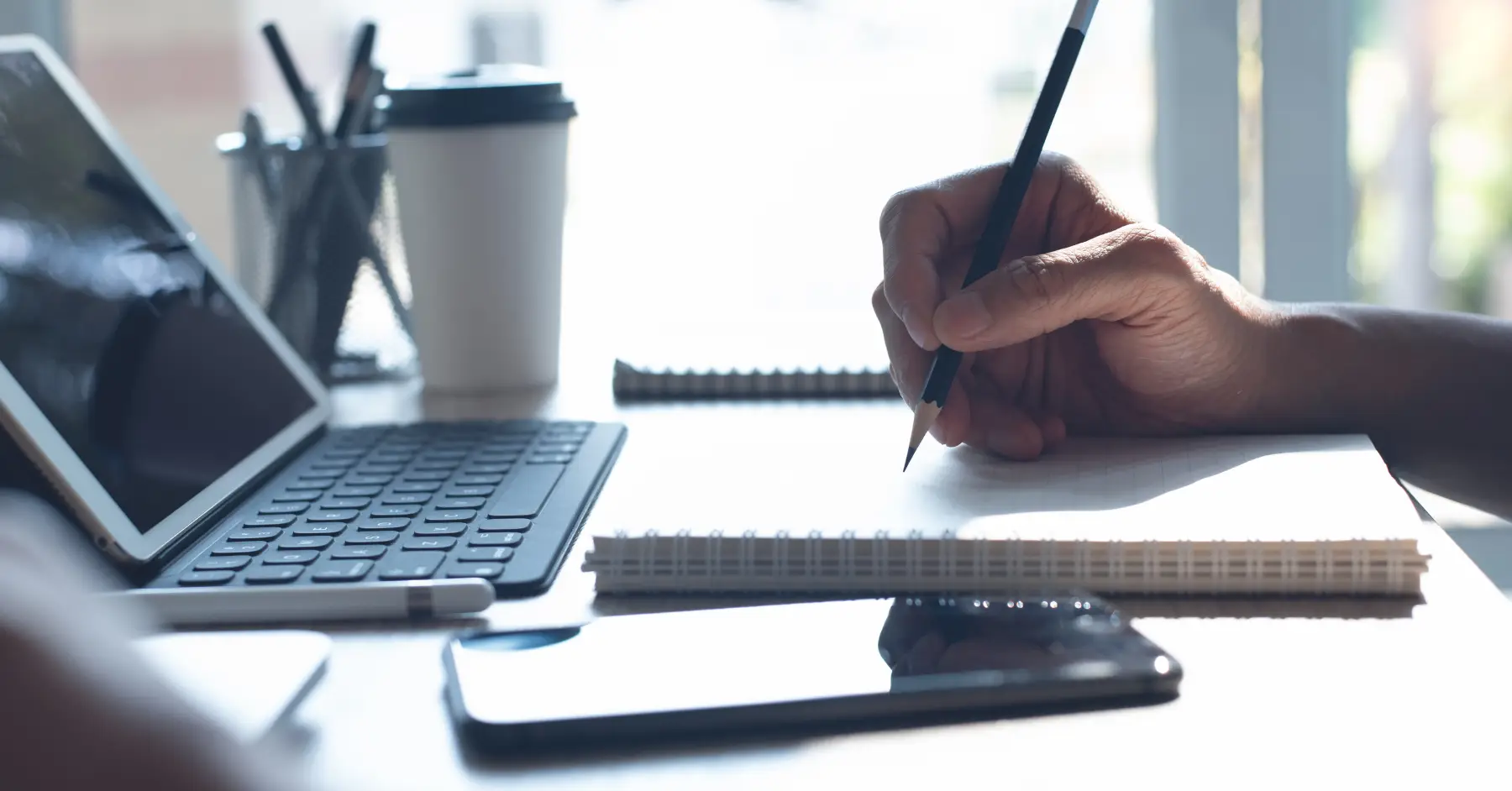 Person writing notes next to a laptop and mobile phone on a desk.