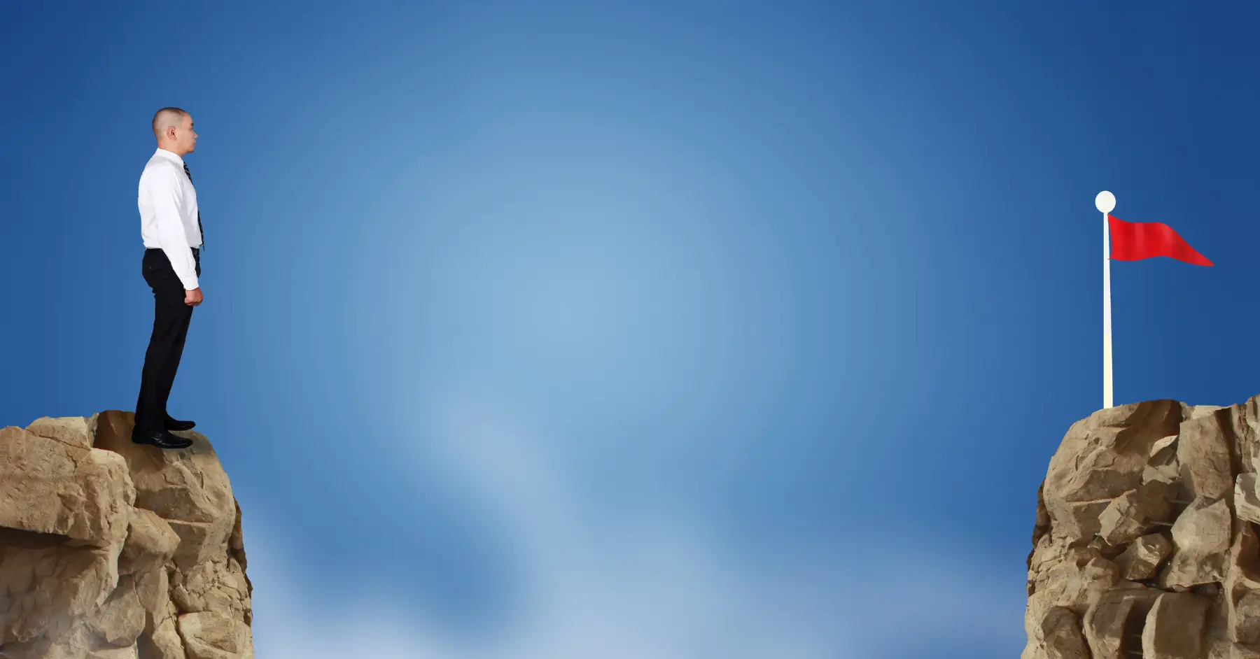 Businessman standing at the edge of a cliff looking towards a flag on another peak.