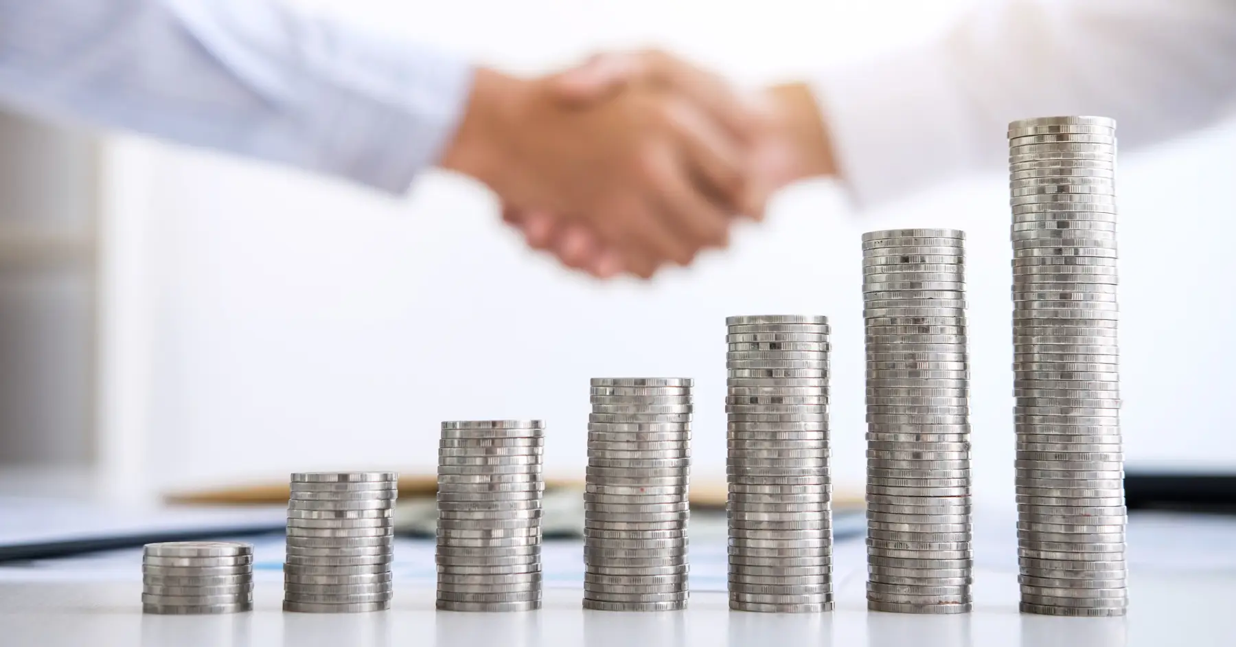 Two professionals shaking hands over a desk, with rising stacks of coins symbolizing successful financial growth and investment in a startup.