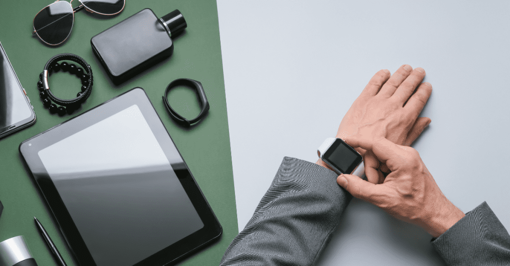 Person adjusting a smartwatch on their wrist, surrounded by various accessories including sunglasses, a tablet, and a smartphone on a green background.