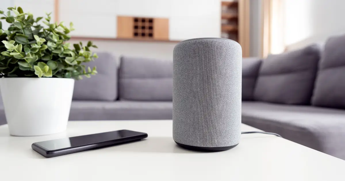 Smart speaker on a living room table alongside a smartphone and a potted plant, illustrating modern smart home technology.