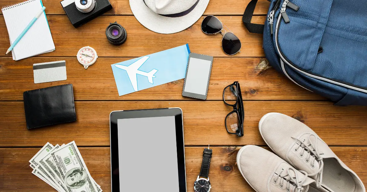 Assorted travel accessories and gadgets spread on a wooden surface, including a backpack, tablet, passport, and sunglasses, ready for a trip.