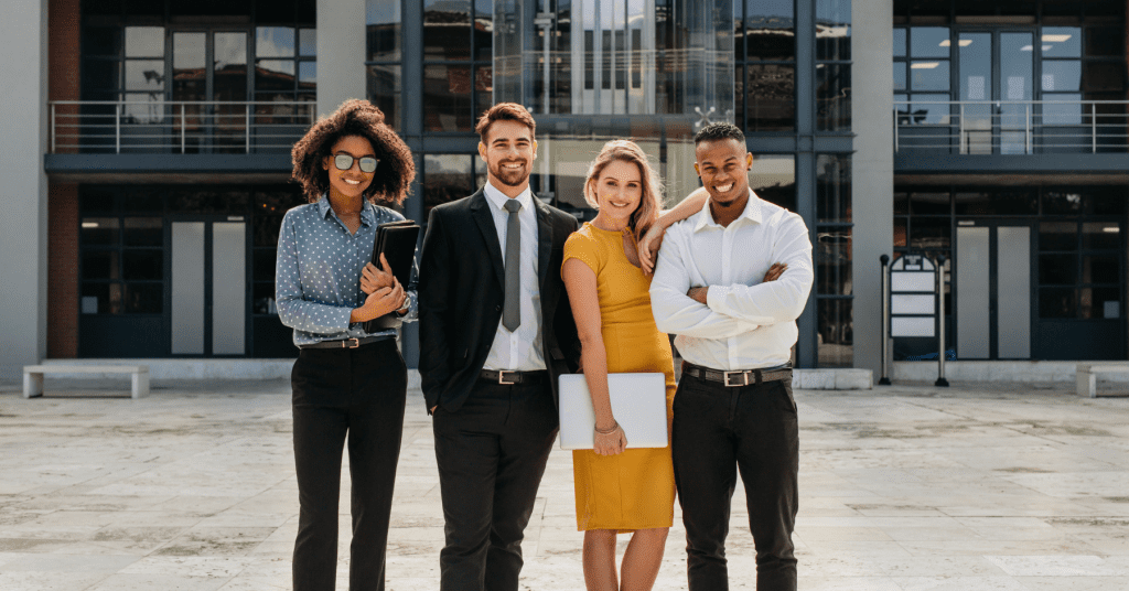 Four diverse young professionals standing confidently in front of a modern office building, smiling at the camera.