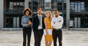 Four diverse young professionals standing confidently in front of a modern office building, smiling at the camera.