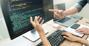 Two people discussing code on a computer screen, pointing at various elements in a programming environment.