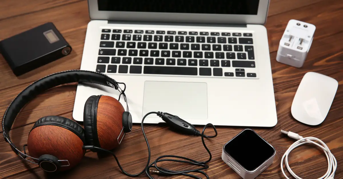 Laptop with assorted accessories including headphones, a portable hard drive, a USB charger, and a mouse on a wooden desk.
