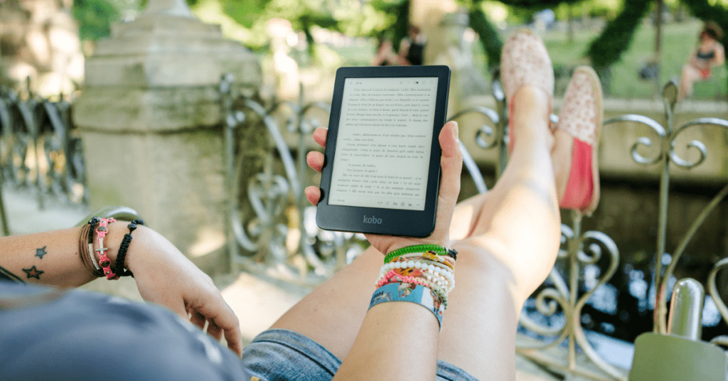 Person relaxing in a park with a Kobo e-reader in hand, showcasing the device's screen filled with text.