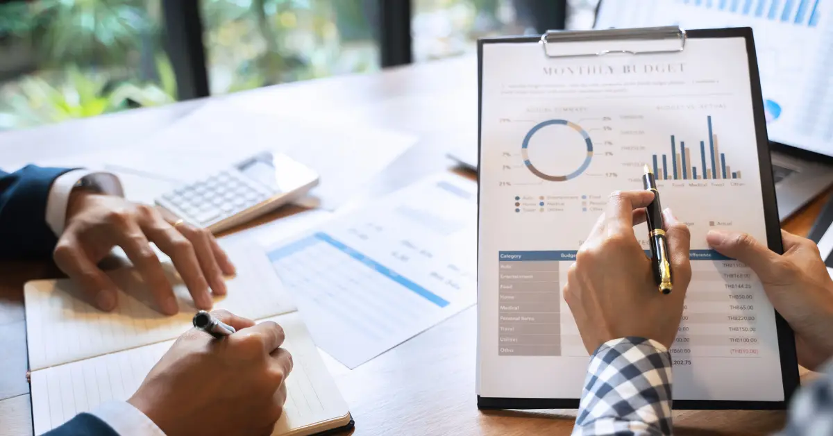 Two professionals reviewing a financial monthly budget report on a clipboard, with focus on budget analysis charts and a calculator on the desk.