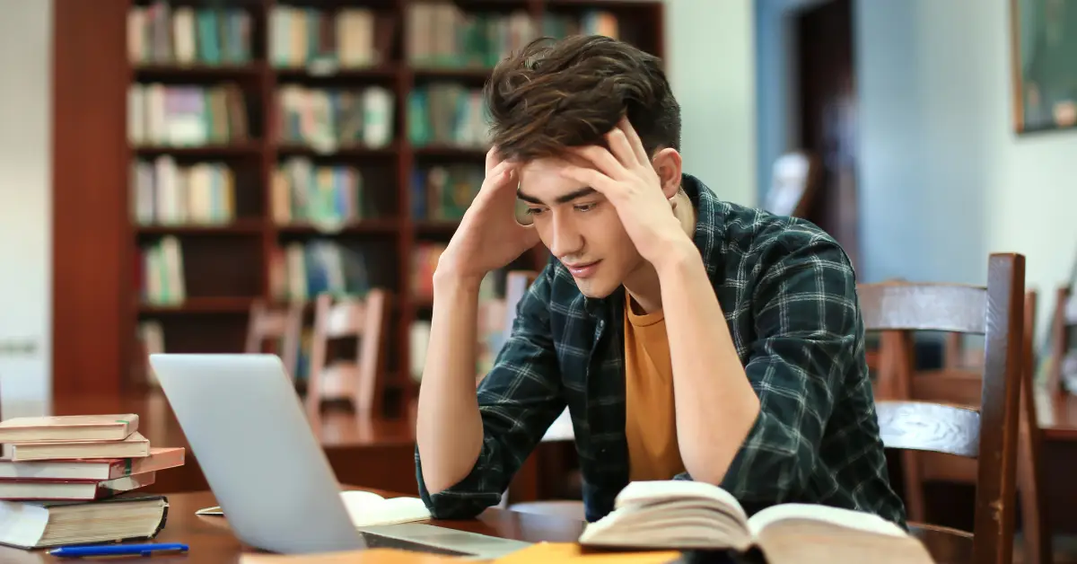 Stressed male student studying in a library with laptop and books
