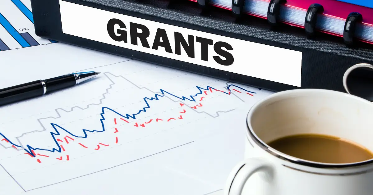 A folder labeled 'GRANTS' with financial charts and a coffee cup, symbolizing the pursuit of funding opportunities for technology startups.