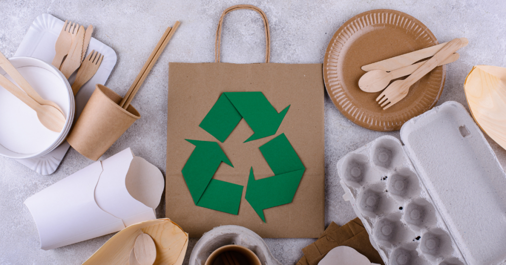 Assorted eco-friendly packaging materials including compostable utensils and recycled paper products with a recycling symbol.