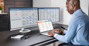 Professional technical project manager working on a laptop and large monitor displaying project management tools and calendars.