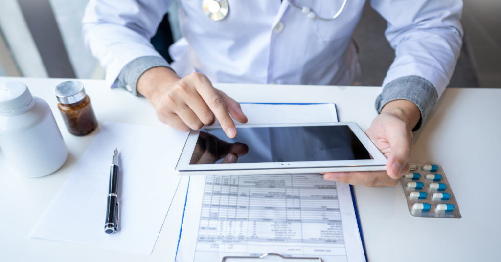 Doctor using a tablet to review patient information with medical records and medication visible on the table.